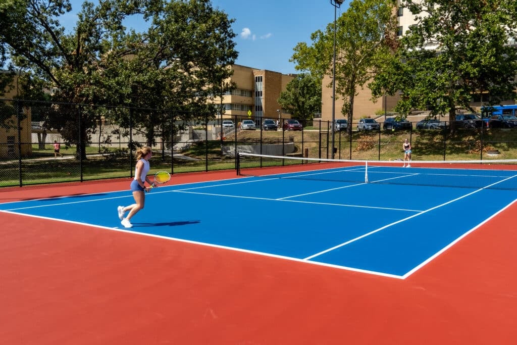 A young woman prepares to hit a tennis ball on a vibrant blue and red court, with trees and buildings in the background.