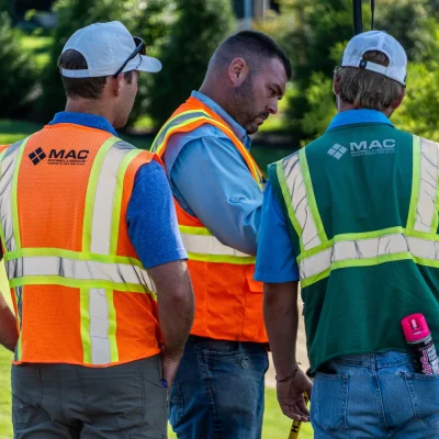 Three construction workers wearing reflective safety vests, two orange and one green with "MAC" logos, stand outdoors on a sunny day.